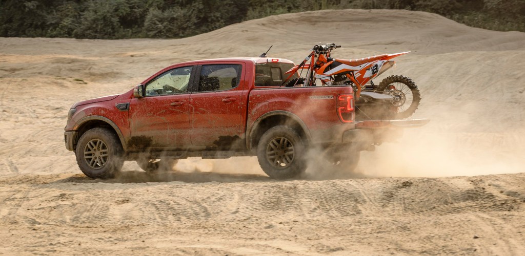 Red Ford Ranger midsize pickup truck off-roading across sand, a dirt bike in its bed.