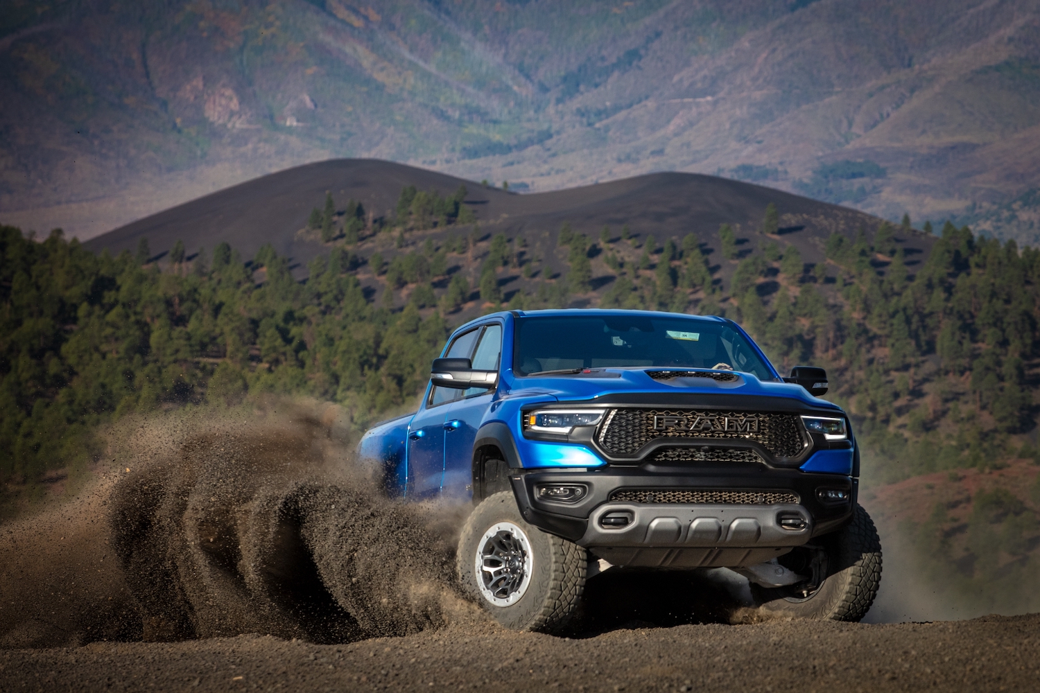 A blue Ram 1500 pickup truck showing off its TRX Hellcat supertruck engine by skidding across the dirt, with wooded hills in the background.
