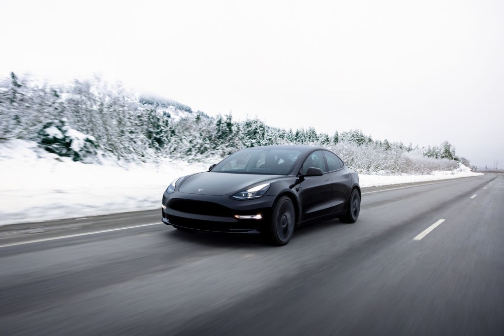 A 2022 Tesla Model 3 electric luxury sedan model in black driving down a highway near a winter forest