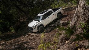White Toyota 4x4 pickup truck showing off its TRD Pro hardware by traversing a steep rock face in a wooded off-road trail.