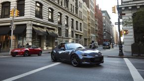 A front view of a grey 2022 Mazda Miata RF and a red Miata convertible on a city street with buildings in the background.