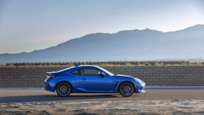 Side profile of a blue 2022 Subaru BRZ coupe, trim level uncertain, parked on a dirt road against the backdrop of sprawling mountains