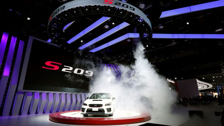 A white 2019 Subaru S209 STI sports car sits on a stage at an automotive show, with manufactured fog floating around it for dramatic effect