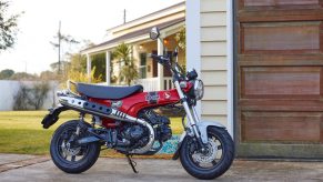 A red-and-black 2023 Honda ST125 Dax next to a garage