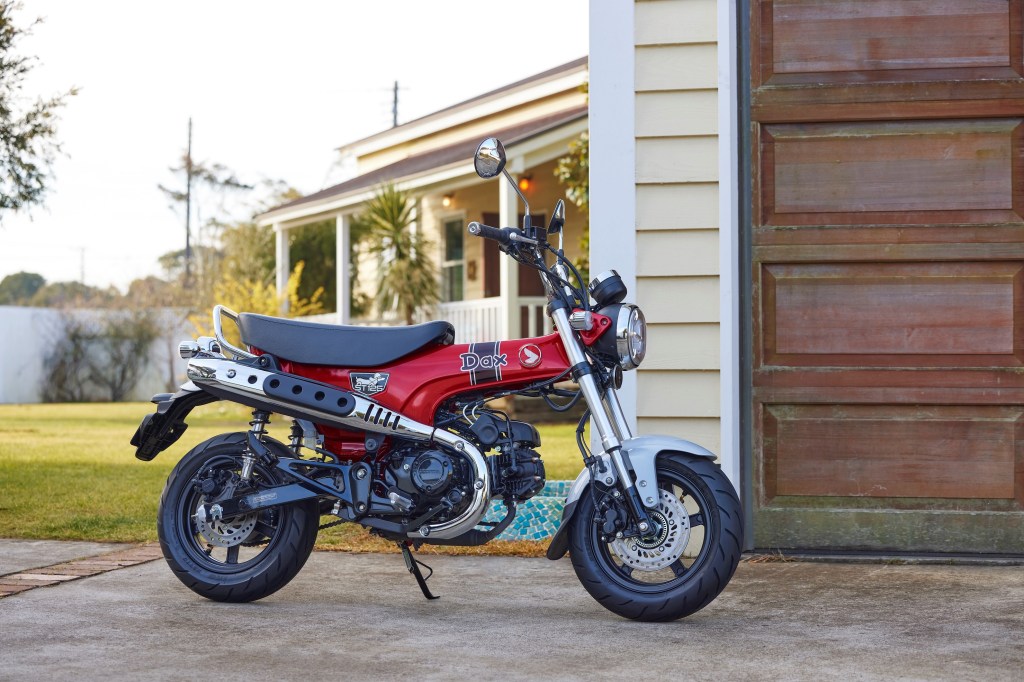 A red-and-black 2023 Honda ST125 Dax next to a garage