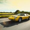 A 3/4 front view of a yellow Lamborghini Miura driving on a road with trees and hills in the background.