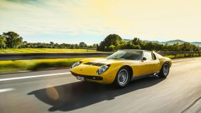 A 3/4 front view of a yellow Lamborghini Miura driving on a road with trees and hills in the background.