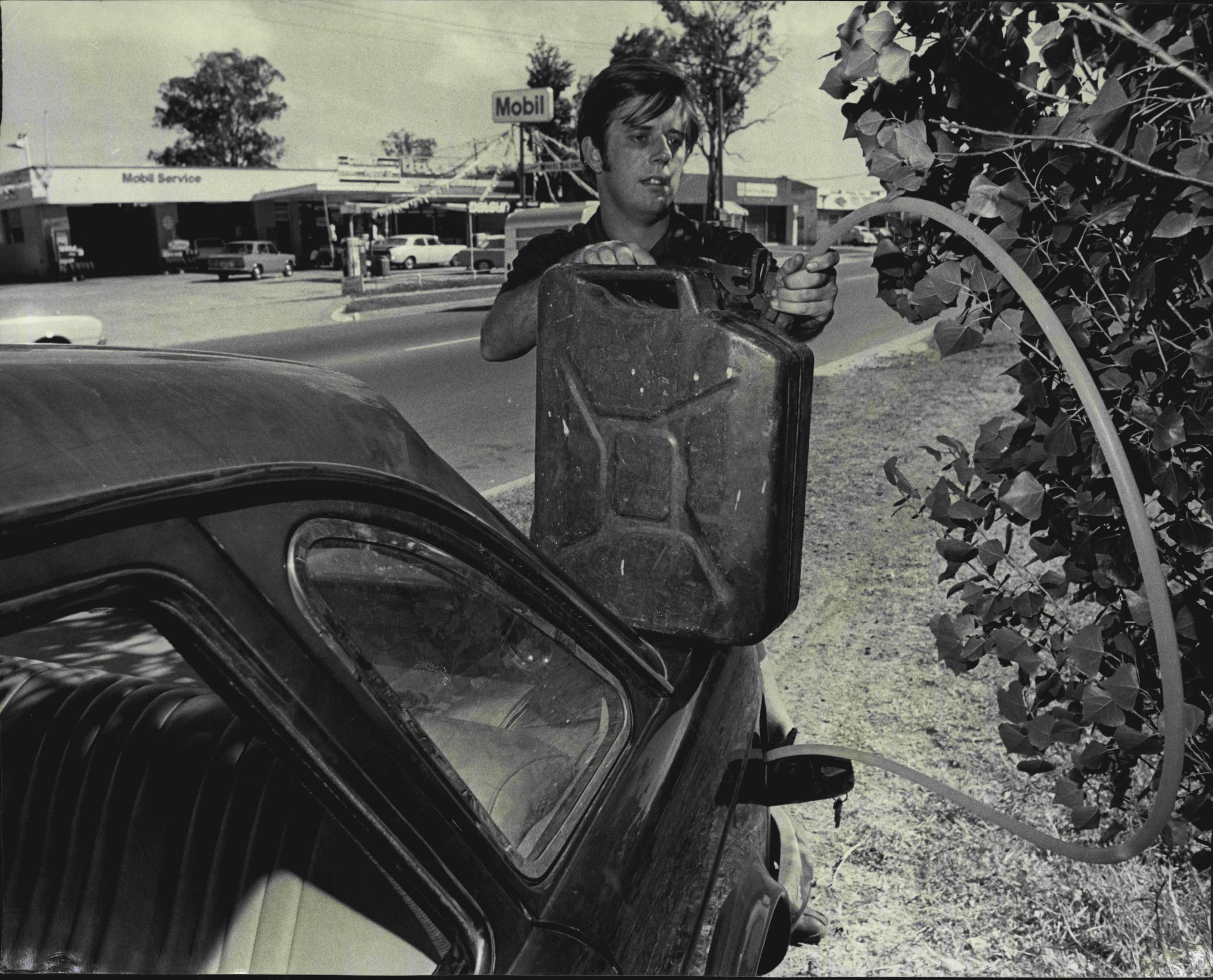 A man refills his gas tank via a jerry can by a station in 1973