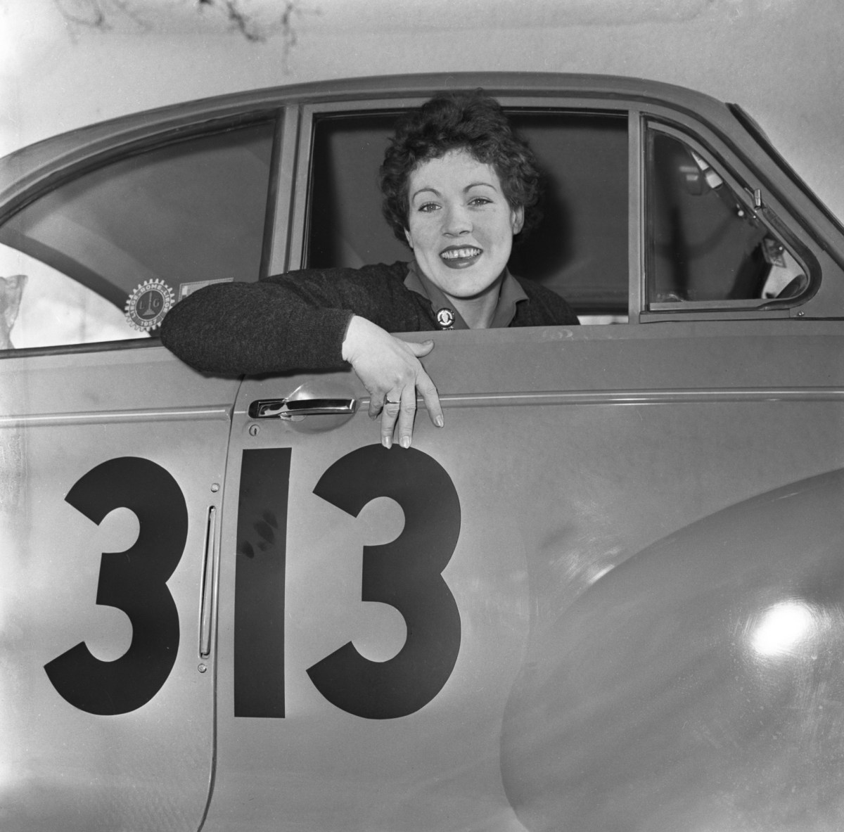 A black and white photo of rally driver Pat Moss smiling through the window of her car before the start of the 1958 Monte-Carlo Rally.