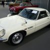 A 3/4 front/profile view of a white 1970 Mazda Cosmo 110S sports car. The car is parked in line with a red sports car at the Grand Palais in Paris, France.