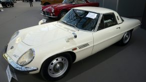A 3/4 front/profile view of a white 1970 Mazda Cosmo 110S sports car. The car is parked in line with a red sports car at the Grand Palais in Paris, France.