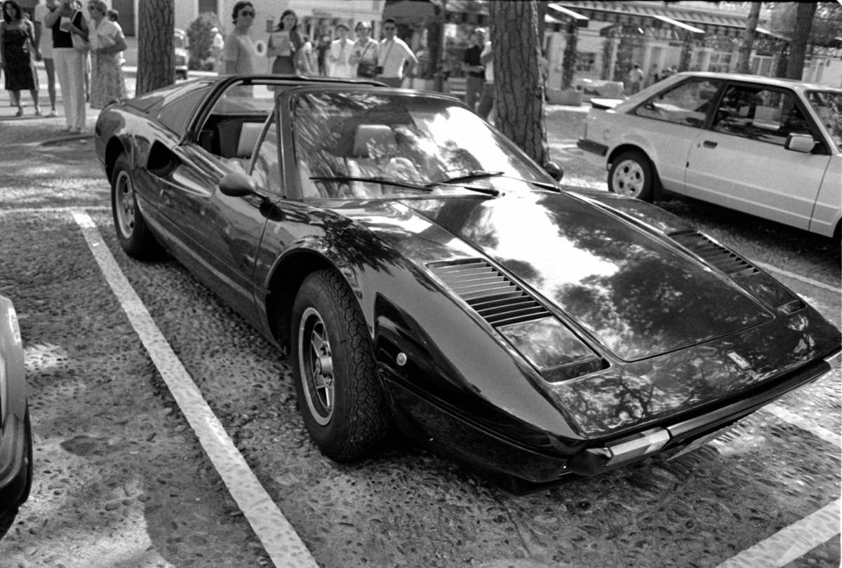 A black and white image of a 1982 Ferrari 308 GTB parked on a street in Monaco with people in the background.