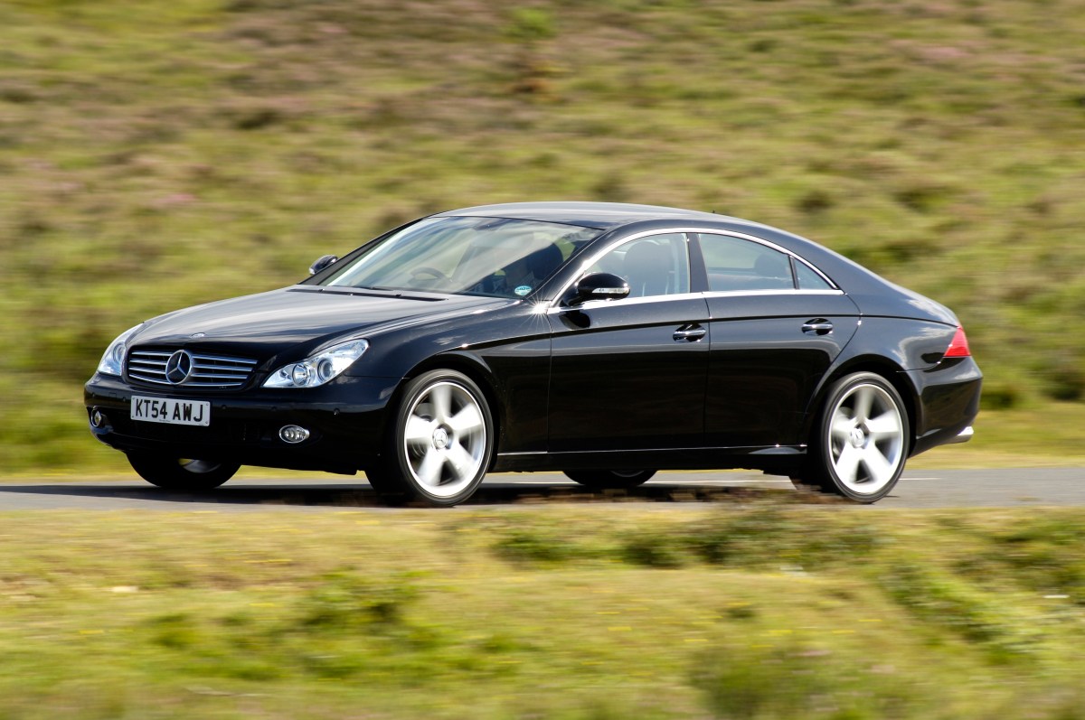 A semi-profile view of a black 2005 Mercedes-Benz CLS500 driving on a road with a grassy hill in the background.