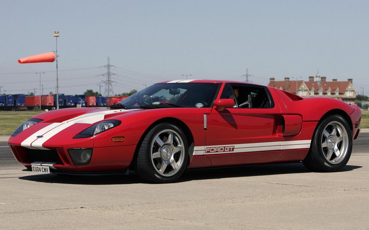 A profile view of a red Ford GT supercar on a runway. A windsock and buildings can be seen in the background.