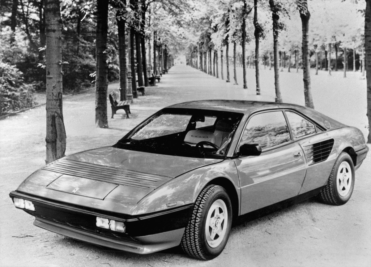 A black and white 3/4 front view of a 1980 Ferrari Mondial 8 parked on a tree-lined walkway.