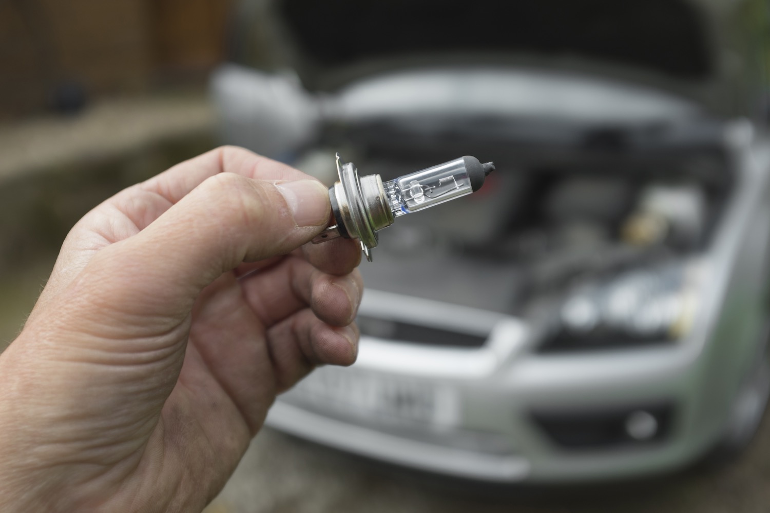 A hand holding a headlight replacement bulb in front of a car with its hood up.