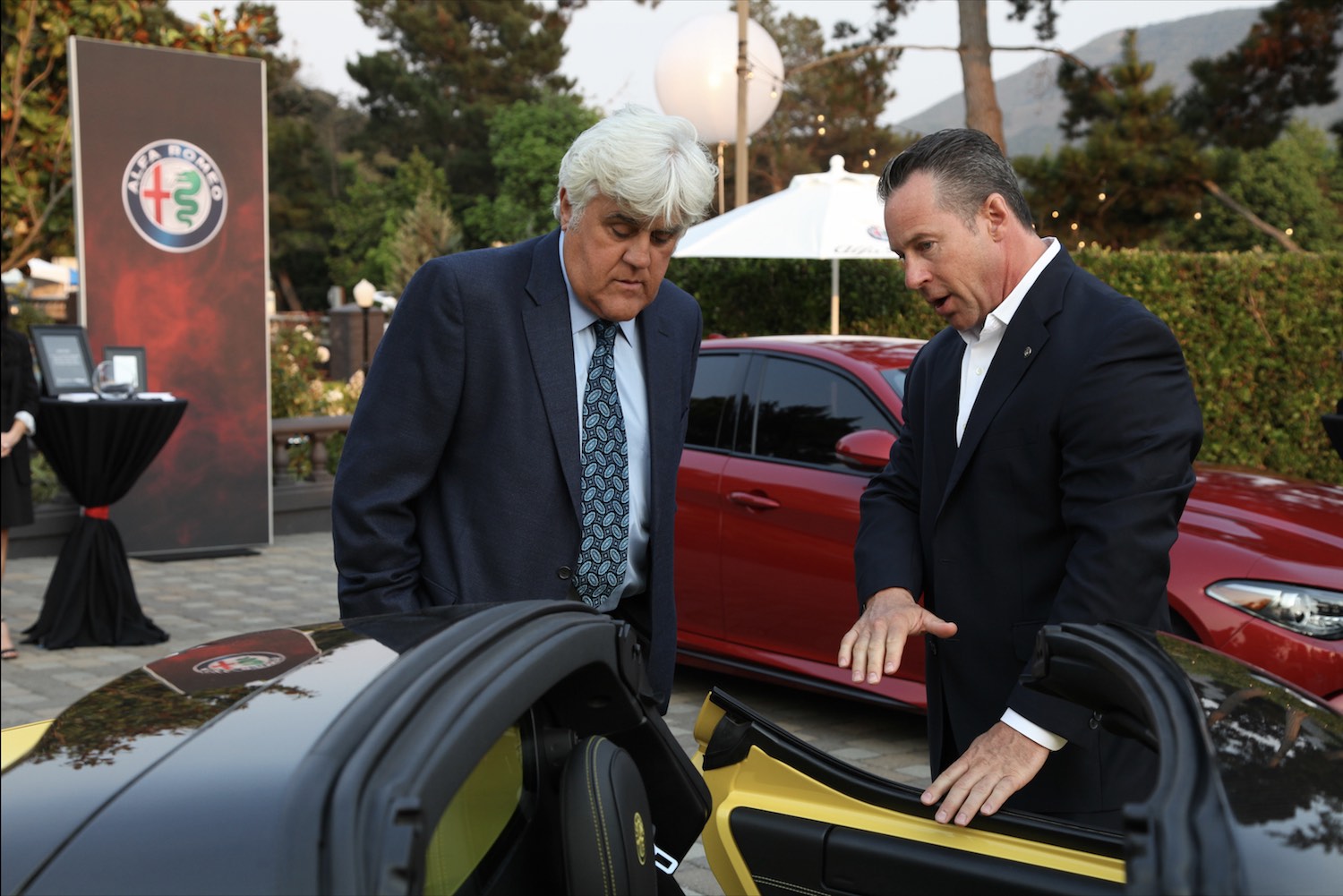 Jay Leno listening to the dealership pitch next to a yellow Alfa Romeo.
