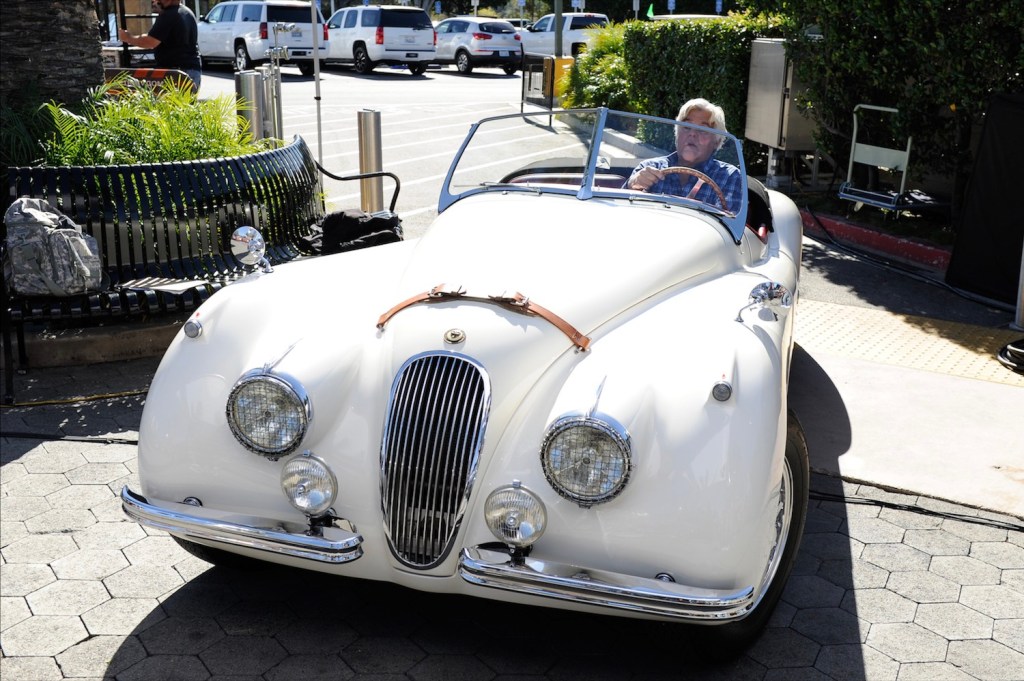 Jay Leno steering a white Jaguar convertible into a driveway.