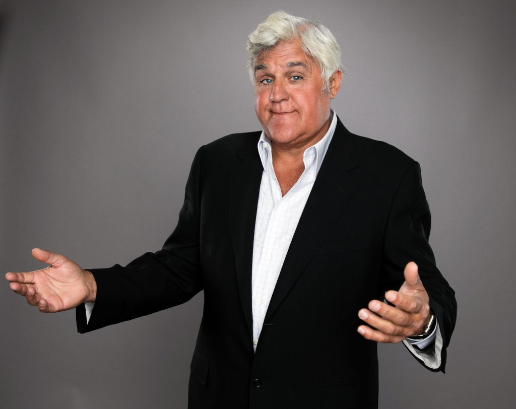 Jay Leno shrugs and smiles, wearing a suit, in a publicity photo.