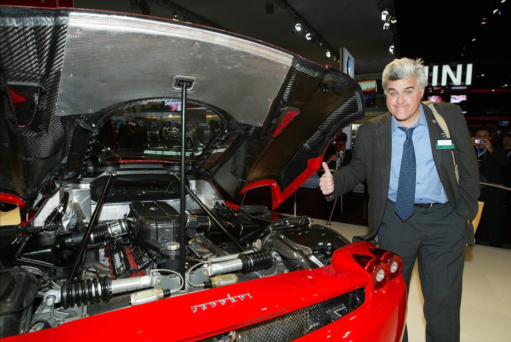 Jay Leno next to a Ferrari sportscar at an auto show.