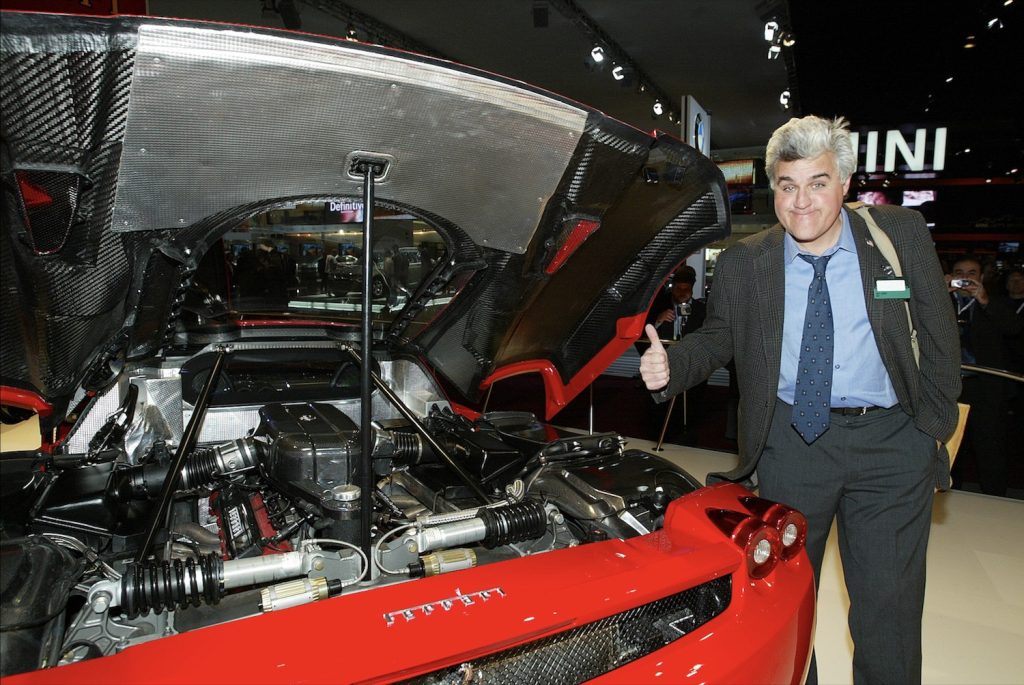 Jay Leno gives the thumbs up next to a red Ferrari Enzo sports car at a crowded auto show.