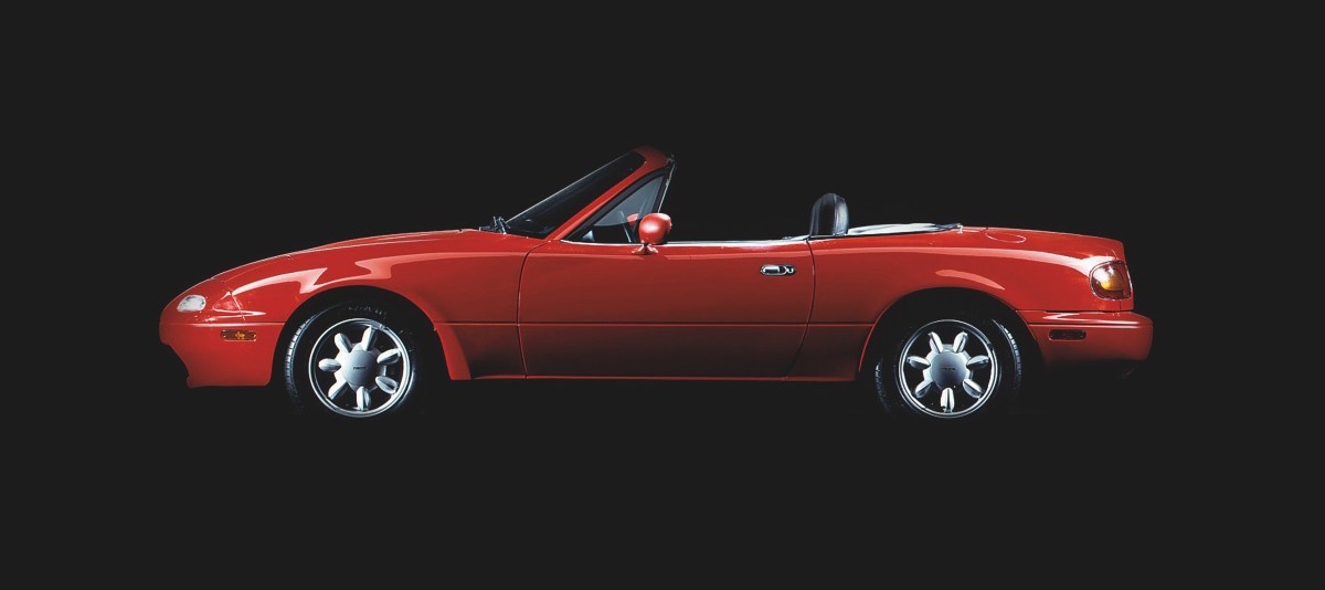 A profile view of a red 1990 Mazda MX-5 Miata with a black studio background.