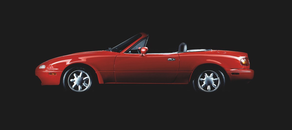 A profile view of a red 1990 Mazda MX-5 Miata with a black studio background.