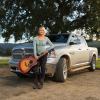 Woman with a guitar leans against a Ram 1500 parked on a farm, in front of a tree.