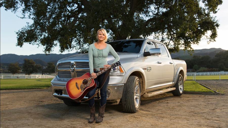 Woman with a guitar leans against a Ram 1500 parked on a farm, in front of a tree.