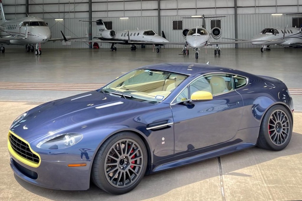 A blue-and-yellow modified 2006 Aston Martin V8 Vantage parked by an airplane hanger