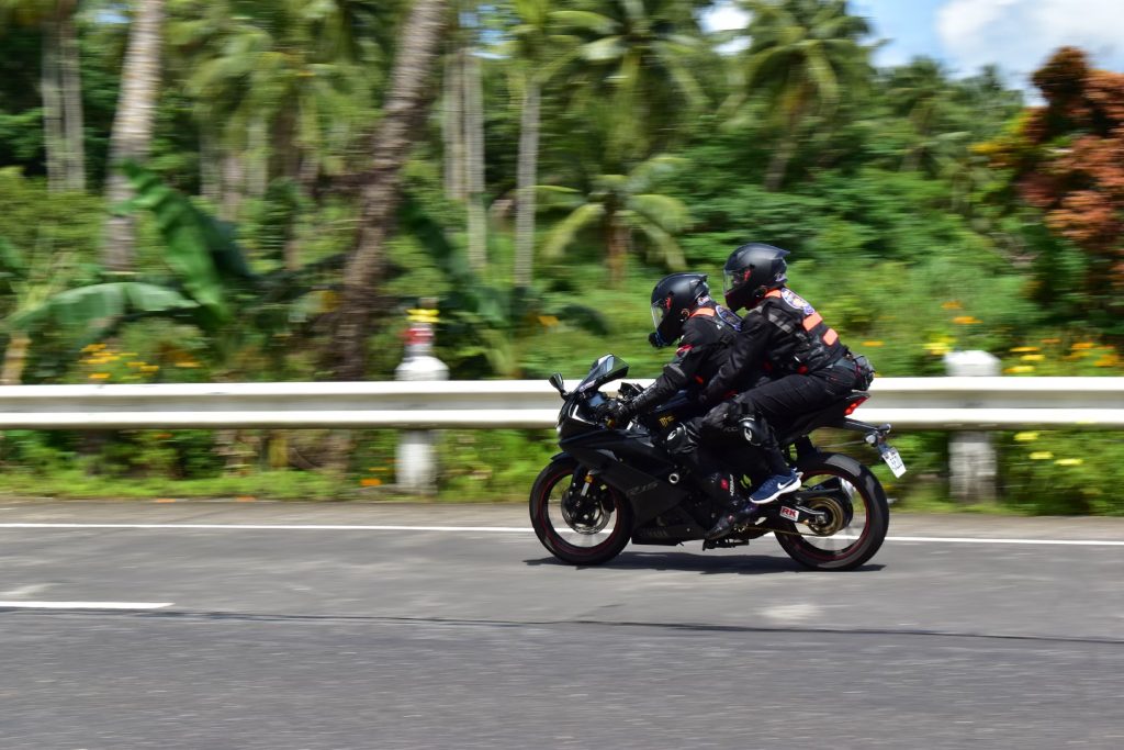 A safety-gear-clad motorcycle rider and passenger ride a sportbike on a jungle road