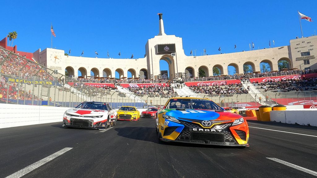 Pack of NASCAR Next Gen cars racing towards the camera, crowded grandstands in the background.