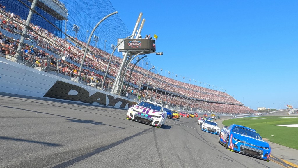 A row of NASCAR Next Gen cars racing around the tri-oval during the Daytona 500, with packed stands visible in the background.