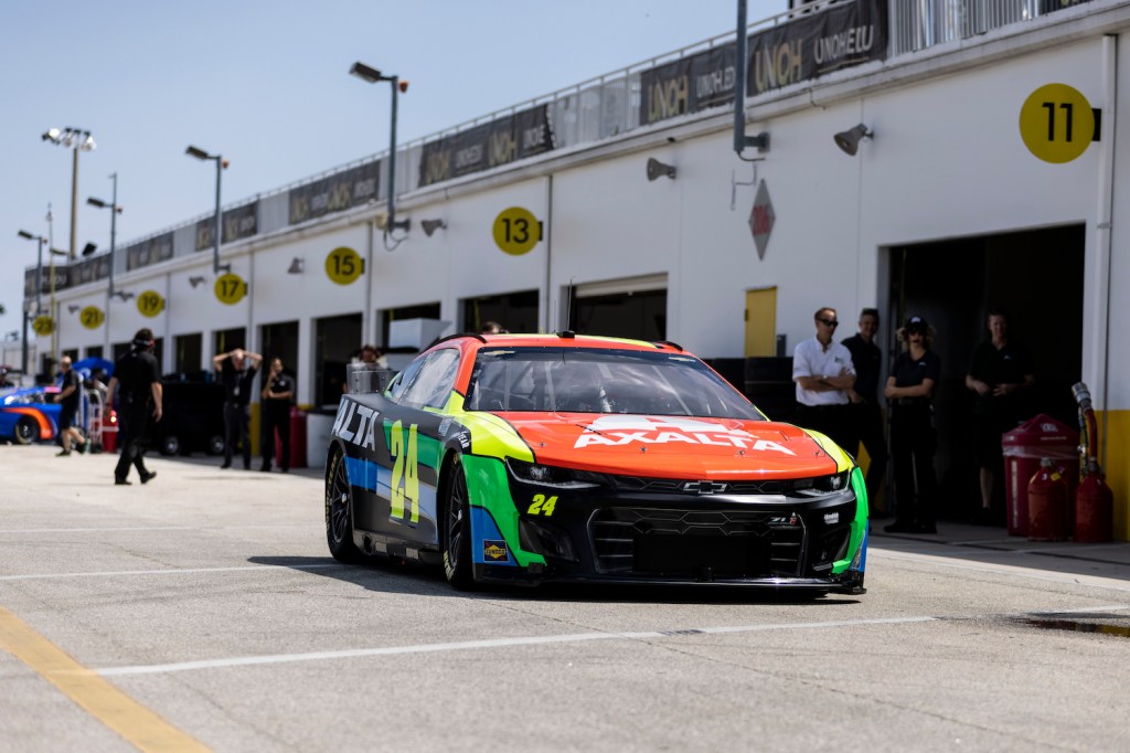 A NASCAR Next Gen race car parked in pit road, technicians standing in the background.