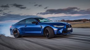 A blue BMW M4 Competition powersliding through a corner of a race track with smoke coming from the rear wheels.
