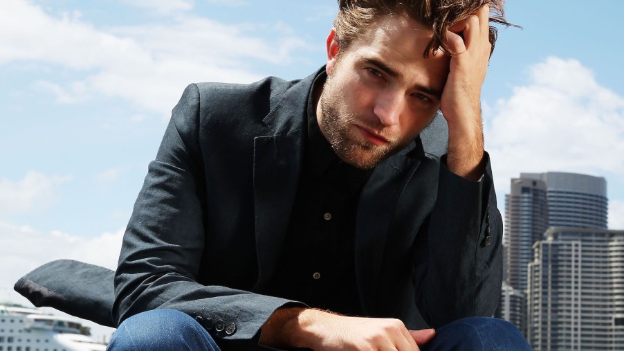 Publicity shot of actor Robert Pattinson dressed in jeans and a suit jacket, the daytime skyline of a city visible behind him.