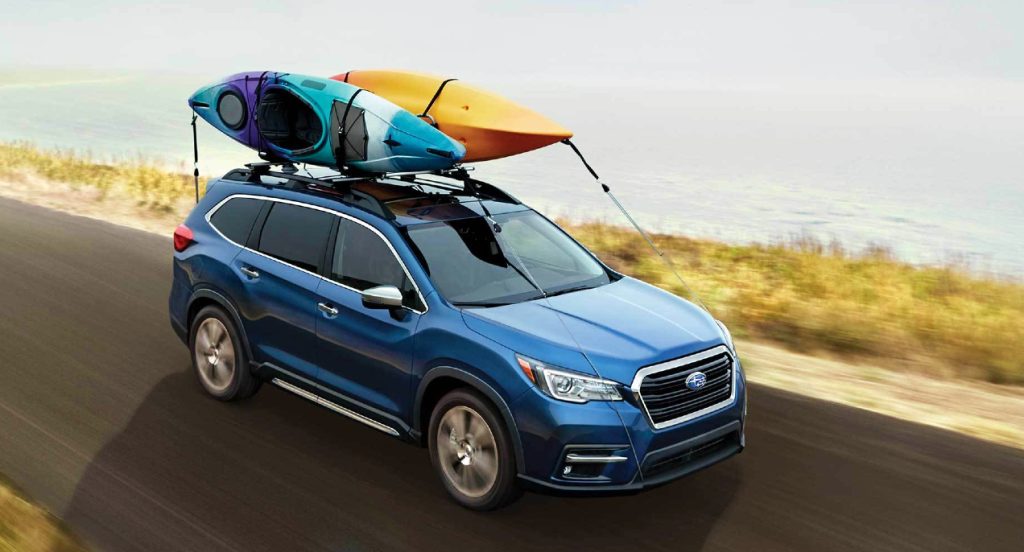 A blue 2022 Subaru Ascent mid-size SUV is driving on the road with kayaks strapped to its roof. 