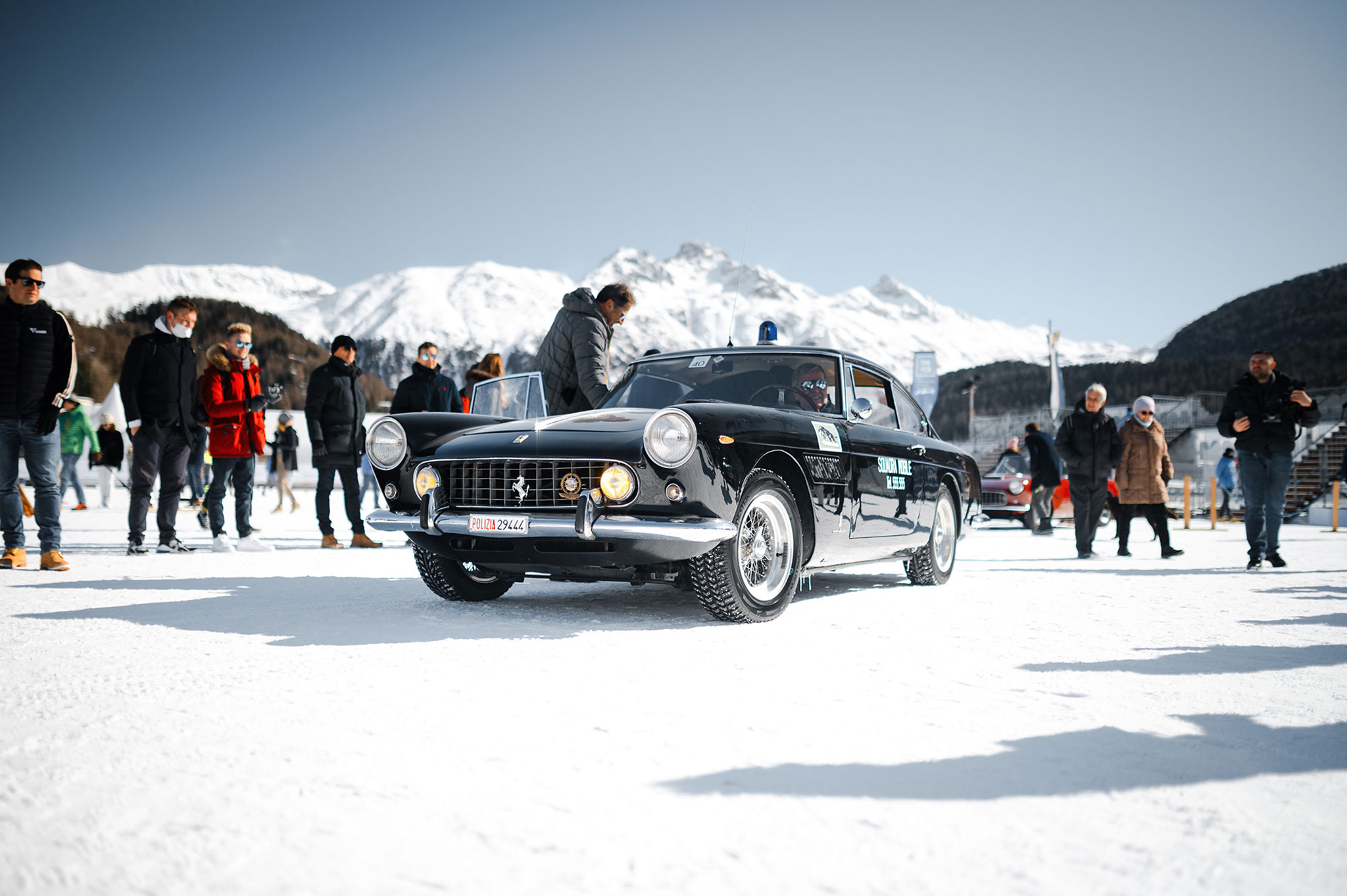 A 3/4 front view of a black Ferrari 250 GT Berlinetta parked on the frozen lake of St. Moritz with people and mountains in the background.