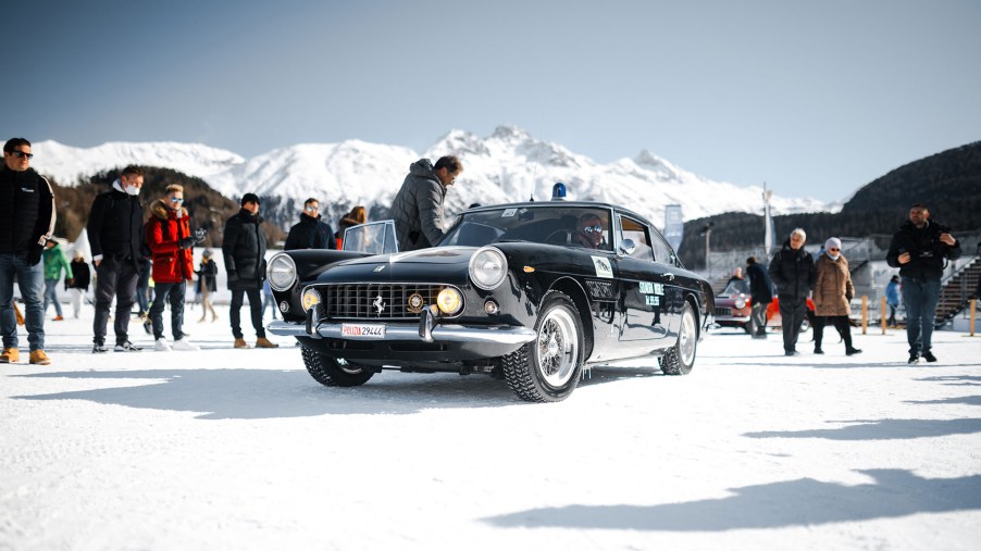A 3/4 front view of a black Ferrari 250 GT Berlinetta parked on the frozen lake of St. Moritz with people and mountains in the background.