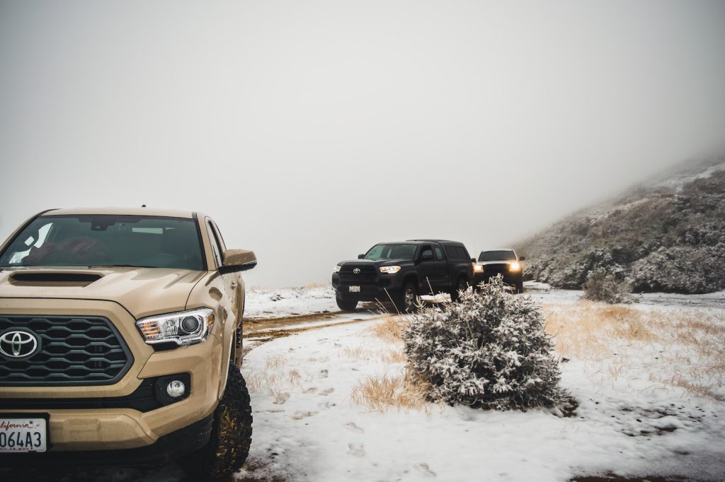 A convoy of third, second, and first generation Toyota Tacoma trucks off-roading on a snowy, mountain trail.