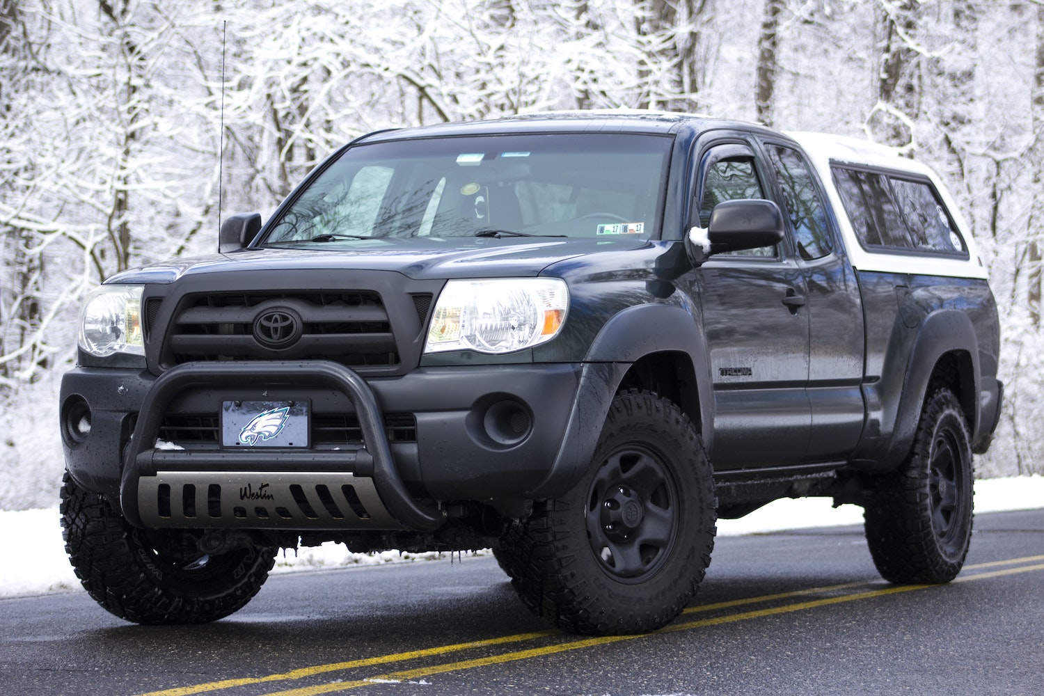 Black first generation Toyota Tacoma truck parked on a road in front of snow-covered trees.