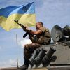 Ukrainian military soldier holds a machine gune and smiles while sitting atop an armored vehicle flying a Ukraine flag.