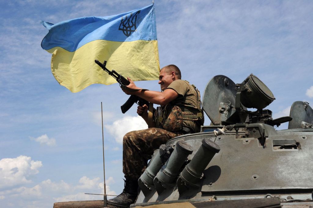Ukrainian military soldier holds a machine gune and smiles while sitting atop an armored vehicle flying a Ukraine flag.