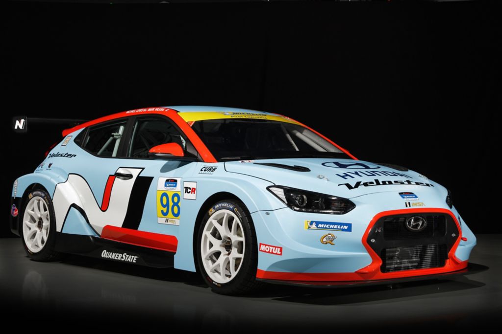 A 3/4 front view of a light blue and red Hyundai Veloster N TCR racecar in a black studio background.