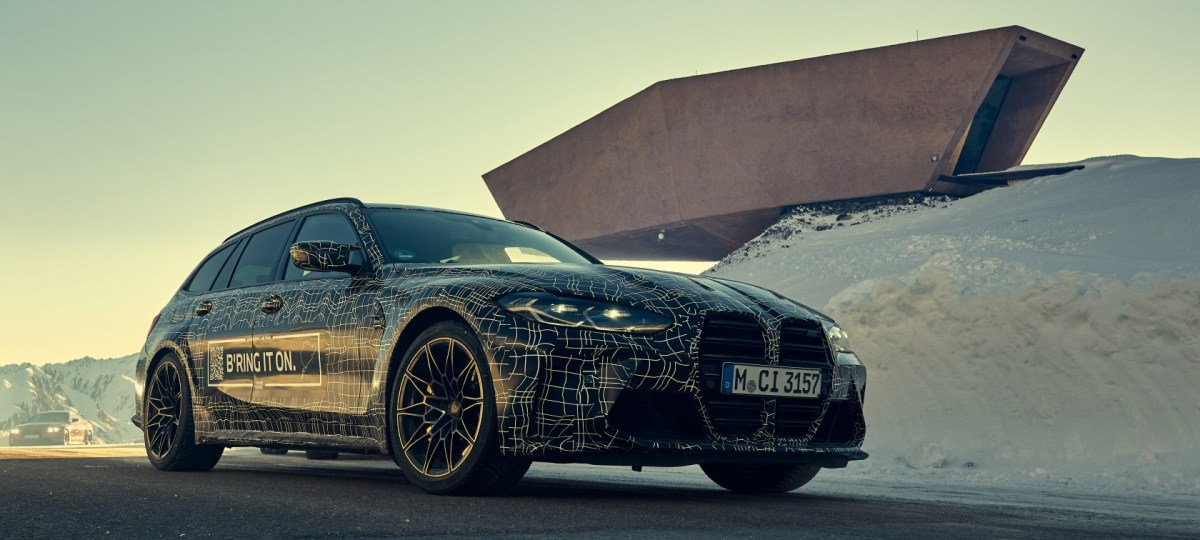 A 3/4 front view of a camouflaged BMW M3 Touring parked in front of a modern building. 