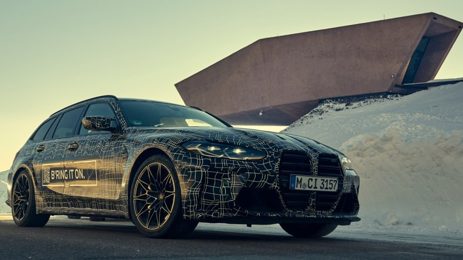 A 3/4 front view of a camouflaged BMW M3 Touring parked in front of a modern building.