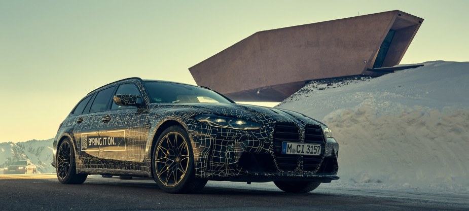 A 3/4 front view of a camouflaged BMW M3 Touring parked in front of a modern building.