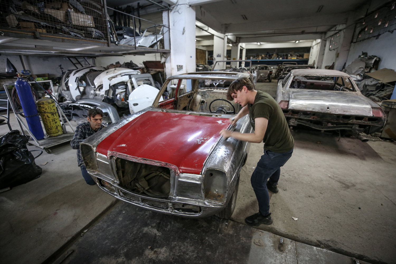 Two men in a crowded auto shop are stripping the paint off a classic car.