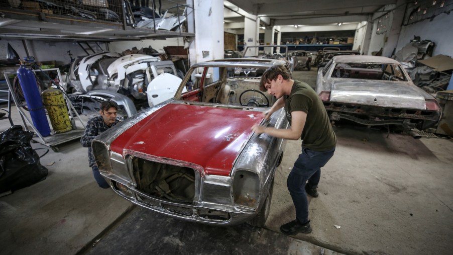 Two men in a crowded auto shop are stripping the paint off a classic car.
