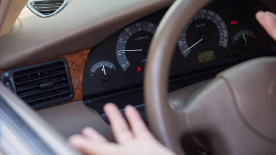 Close up of a half-empty fuel gauge in an older-model-year vehicle; a pair of hands making an exasperated gesture are blurred in the forefront